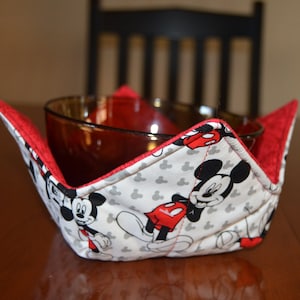 May include: A handmade bowl cozy featuring a Mickey Mouse print. The cozy has a white background with Mickey Mouse illustrations and red accents. It is designed to hold a bowl and protect hands from heat.