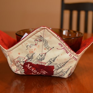 May include: A cream-colored bowl cozy with a red interior, featuring a map design with the word "Hogwarts" on a banner. The cozy is designed to hold a bowl and is placed on a wooden table.