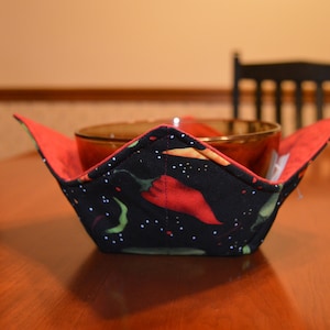 May include: A black bowl cozy with a red chili pepper pattern, holding a brown bowl. The interior is red. The cozy sits on a wooden table, with a black chair in the background.