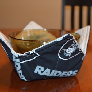 May include: A black and white fabric bowl cozy with the word "RAIDERS" printed on it. The cozy is holding a clear glass bowl.