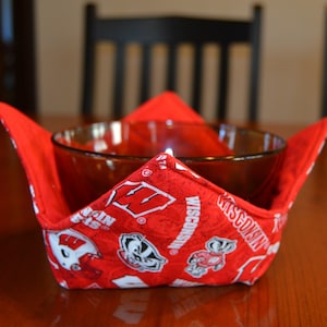 May include: A red bowl cozy with a Wisconsin Badgers theme. The cozy is made of red fabric with white logos and text. A clear glass bowl sits inside the cozy, which is on a wooden table.