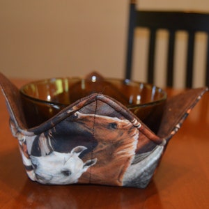 May include: A brown glass bowl sits inside a fabric bowl cozy. The cozy features a horse-themed print with images of white and brown horses. The cozy is designed to hold the bowl and keep its contents warm.