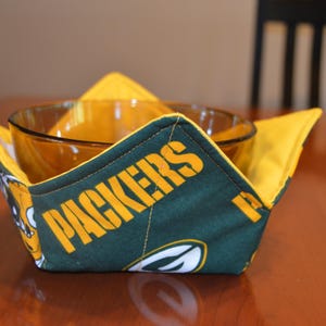 May include: A green and yellow bowl cozy with the word "PACKERS" in yellow lettering. The cozy is holding a clear glass bowl. The cozy is made of fabric and has a yellow interior.