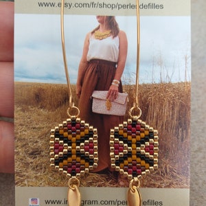 May include: Gold hoop earrings with a square-shaped beaded design in black, red, and gold. The beads are woven together in a geometric pattern. The earrings have a gold teardrop charm hanging from the bottom.