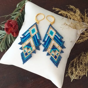 Peut inclure: Une paire de boucles d'oreilles en forme de feuille, faites de perles. Les boucles d'oreilles présentent un motif de perles bleues, blanches et dorées. Elles sont présentées sur un coussin blanc. Fermoirs dorés.