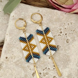 May include: Gold-coloured earrings with a rectangular beaded design. The earrings display a pattern of white, navy blue, and mustard yellow beads. Each earring has a gold-coloured teardrop-shaped charm hanging from the bottom.