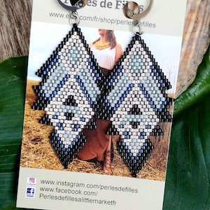 May include: A pair of silver and black geometric beaded earrings with a silver hook fastening. The earrings are shaped like a triangle with a pointed bottom. The beads are arranged in a pattern of black, silver, and light blue. The earrings are displayed on a card with the text "Perles de Filles" and a website address.