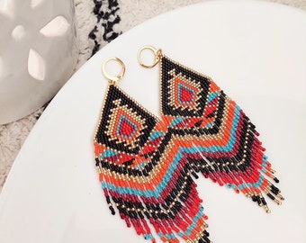 Peut inclure: Une paire de boucles d'oreilles perlées colorées avec un motif géométrique. Les boucles d'oreilles présentent un motif noir, rouge, orange et turquoise avec des accents dorés et des franges. Elles sont présentées sur une surface blanche.