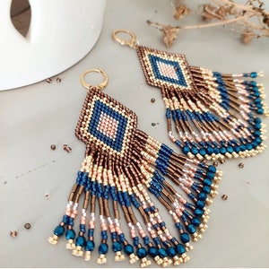 Op de afbeelding: Een paar lange, geperde oorbellen met een geometrische diamantvorm in het midden. De oorbellen zijn gemaakt van bruine, gouden, blauwe en roze kralen.
