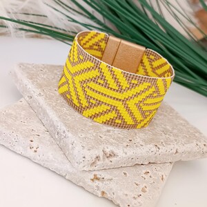 Op de afbeelding: Een geel en bruin kralenarmband met een geometrisch patroon. De armband heeft een magnetische sluiting en wordt getoond op een getextureerd, lichtgekleurd stenen oppervlak. Groen en wit blad is op de achtergrond.
