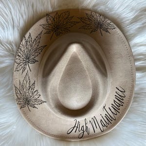 May include: A beige fedora hat with black leaf designs and the text "High Maintenance" written in cursive.