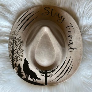 May include: A beige fedora hat with black text that says "Stay feral" and a black silhouette of a wolf howling at the moon in a forest setting.