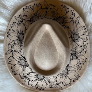 Könnte beinhalten: Beiger Fedora-Hut mit breiter Krempe, verziert mit einem schwarzen Blumenmuster aus Sonnenblumen. Der Hut hat eine tropfenförmige Krone und eine weiche, filzartige Textur, ideal für Sonnenschutz und Mode.