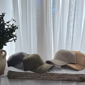 May include: A collection of suede baseball caps in various neutral colors, including gray, olive green, tan, and brown. The caps are displayed on a piece of wood, with a vase of greenery in the background.