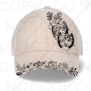 May include: A beige baseball cap featuring a black line art graphic on the front panel and brim. The design displays two horseshoes intertwined with various flowers, leaves, and a butterfly, extending from the side panel onto the curved brim.