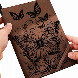 May include: Brown notebook with a detailed black butterfly design. The cover features a large butterfly with intricate wing patterns, surrounded by smaller butterflies and floral accents. A brown elastic band is visible on the right side.
