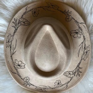 May include: A tan fedora hat with a floral design. The hat features a black floral wreath around the brim. The hat is made of a soft material and has a classic fedora shape.