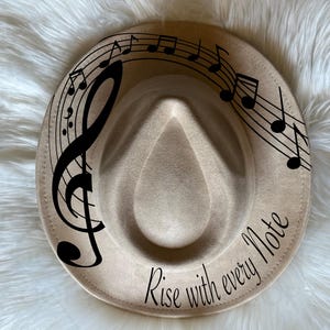 Peut inclure: Un chapeau fedora beige avec un dessin de clé de sol et de notes de musique noires. Le chapeau porte l'inscription "Rise with every note" sur le bord.