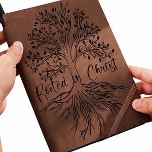 May include: Brown journal with a tree design and the words "Rooted in Christ" in black script. The tree illustration features a detailed trunk, branches, leaves, and roots. The journal has a brown elastic band closure.