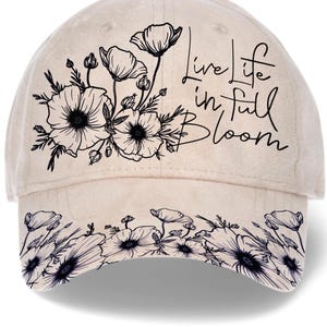May include: A beige baseball cap with a floral design and the text "Live life in full Bloom." The cap features a black floral pattern on the front and brim, with a curved visor and adjustable strap.