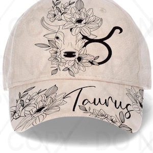 May include: A beige baseball cap featuring a black floral design and the word "Taurus". The design includes a bull's head with flowers and the Taurus zodiac symbol. The cap is a stylish accessory.