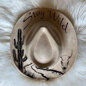 May include: A beige fedora hat with a black design featuring a cactus, a skull, and the text "Stay Wild".