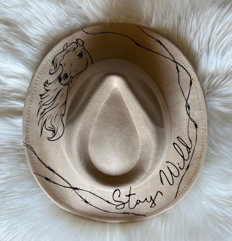 May include: A beige felt fedora hat with a black horse silhouette and the text "Stay Wild" in a cursive font. The hat is decorated with a black barbed wire design.