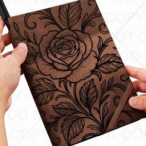 May include: Brown journal with a large rose and leaf design. The rose and leaves are black, and the journal has a brown elastic band. The journal is held open, revealing the inside pages.