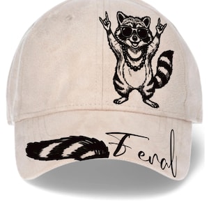 May include: A beige baseball cap featuring a black graphic of a raccoon wearing sunglasses and a beaded necklace while making hand gestures. The word Feral is printed in black script on the brim, accompanied by a black and white striped raccoon tail.