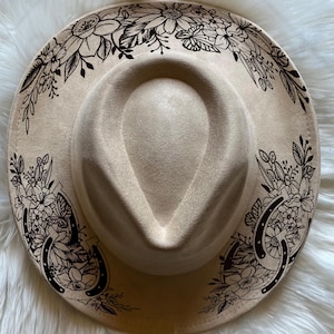 以下が含まれることがあります： ベージュ色のフェドラハット。つばが広く、黒い花柄と蹄鉄のデザインが施されています。涙型のクラウンと柔らかいスエードのような質感が特徴です。どんな服装にもボヘミアンな雰囲気を添える、おしゃれなアクセサリーです。