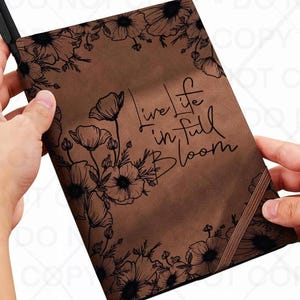 May include: A brown journal with a floral design and the words "Live life in full bloom." The journal features black line art of flowers around the edges and a brown elastic band closure. The journal is held by two hands.