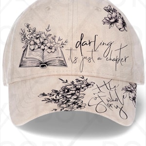May include: Beige baseball cap with floral and book graphics. The cap features the text "darling it's just a chapter, not the whole story" in black script. The cap has a curved brim and a button on top.