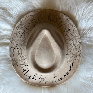 Op de afbeelding: Een beige fedora-hoed met een zwart bladontwerp rond de rand en de tekst "High Maintenance" in cursief.