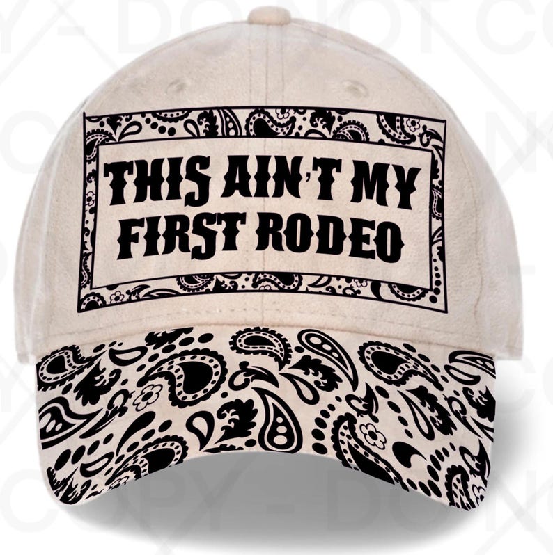 May include: A beige baseball cap with a black paisley pattern on the brim and a rectangular design on the front. The text "THIS AIN'T MY FIRST RODEO" is printed in black, bold letters within the rectangle.