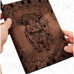 May include: A brown rectangular notebook featuring a dark brown line art illustration of a Highland cow centered on the front cover, surrounded by a decorative border of flowers and leaves that frame the central animal figure on all four sides.