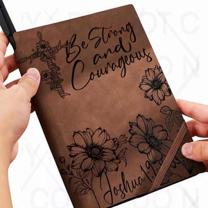 May include: Brown journal with the text "Be Strong and Courageous" and "Joshua 1:9". The journal features floral and cross designs. A black pen is inserted into the journal's spine.