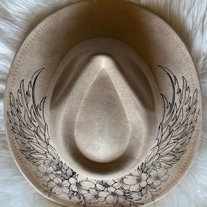 Puede incluir: Un sombrero fedora beige con un diseño de alas y flores negras. El sombrero tiene un ala ancha y una corona estructurada. El diseño de alas está en ambos lados, con un patrón floral a lo largo del ala.