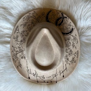 May include: A beige fedora-style hat with a wide brim. The hat features black floral and horseshoe designs, with the text "Wild hearts can't be tamed" in a script font. The hat is displayed on a white, fluffy surface.