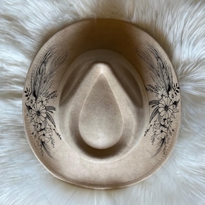 May include: A beige fedora hat with a wide brim. The hat features a floral and feather design in black ink on both sides of the crown. The hat is made of a soft material.