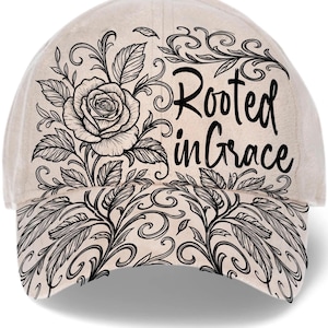 May include: A beige baseball cap featuring a black floral design with a rose and the words "Rooted in Grace." The design includes leaves and swirling patterns, creating an elegant and detailed aesthetic. The cap has a curved brim and an adjustable strap.