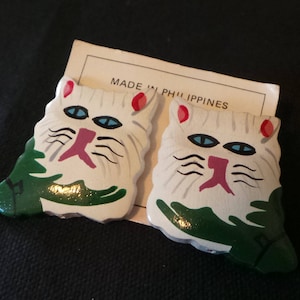 May include: Pair of white ceramic cat earrings with green and red details. The earrings are made in the Philippines.