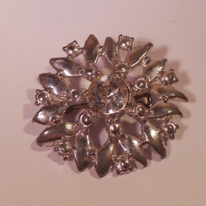 May include: A silver brooch with a large clear rhinestone in the center surrounded by smaller rhinestones and silver leaves.