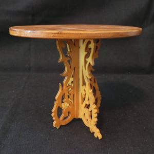 May include: A small, round wooden table with a decorative, intricately carved base. The table is made of light brown wood and has a natural finish.