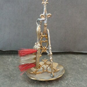 May include: A miniature gold-colored metal stand with a red and white brush, a dustpan, and a decorative top. The stand is on a gray surface.