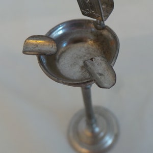 May include: A silver metal ashtray with a tall stand and a round base. The ashtray has a small lip around the edge and a small, curved, metal piece on the side.