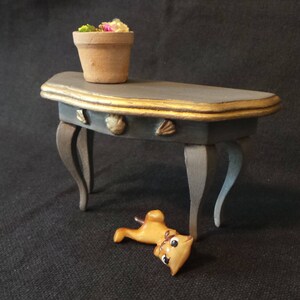 May include: A small, gray, half-moon shaped table with gold trim and decorative seashells. The table has three legs and is made of wood. A small, orange, ceramic cat figurine is lying on the floor in front of the table.