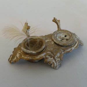 May include: A gold-colored metal decorative stand with two small round depressions. One depression has a small bird's nest with a feather inside. The other depression has a button with three holes.