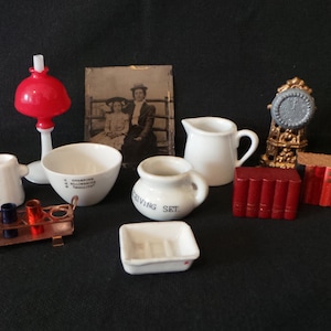 May include: A miniature dollhouse set with a red lamp, white ceramic dishes, a small pitcher, a clock, and books. The set also includes a framed photo of two women.