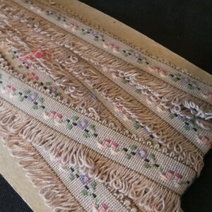 Puede incluir: Primer plano de una cinta beige con un patr&oacute;n floral y flecos. La cinta est&aacute; doblada y apilada sobre una superficie marr&oacute;n.