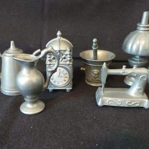 May include: A set of six miniature pewter figurines, including a teapot, a pitcher, a clock, a mortar and pestle, a sewing machine, and a lamp. The figurines are all silver in color and have intricate details.